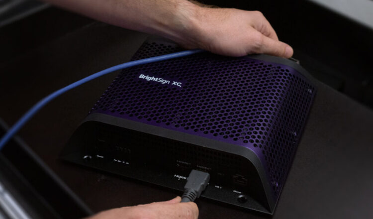 Two hands hold a BrightSign XC5 digital signage player being plugged in, showing easily installation and set up