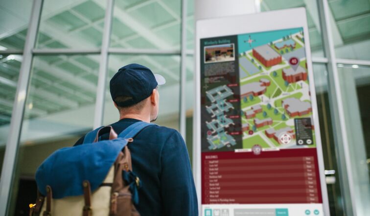 A man with a backpack and baseball cap stands inside a building facing a digital wayfinding kiosk with a campus map on it