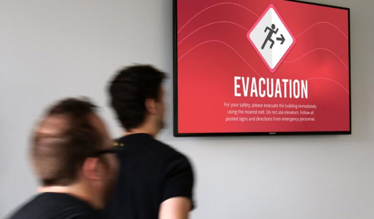 Two men are blurred slightly implying motion as they walk by an emergency alert message on a wall-mounted digital screen