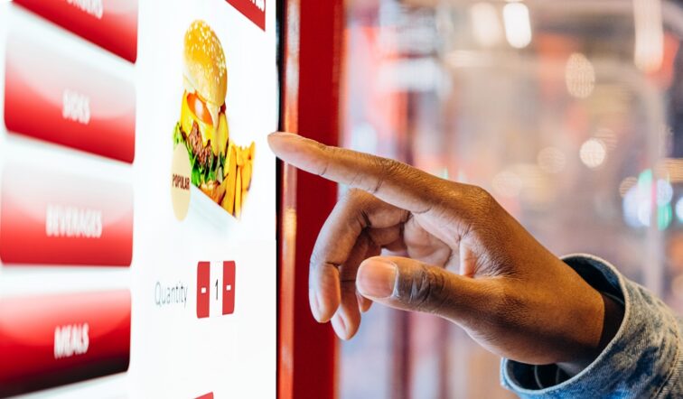 Primer plano de una mano tendida hacia un tablero de menú digital con pantalla táctil en el que se muestra un menú de hamburguesa.
