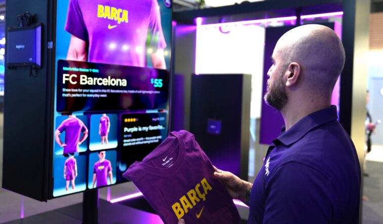 A man lifts a Barca jersey off of a retail display while looking at digital signage with more information about the jersey he picked up